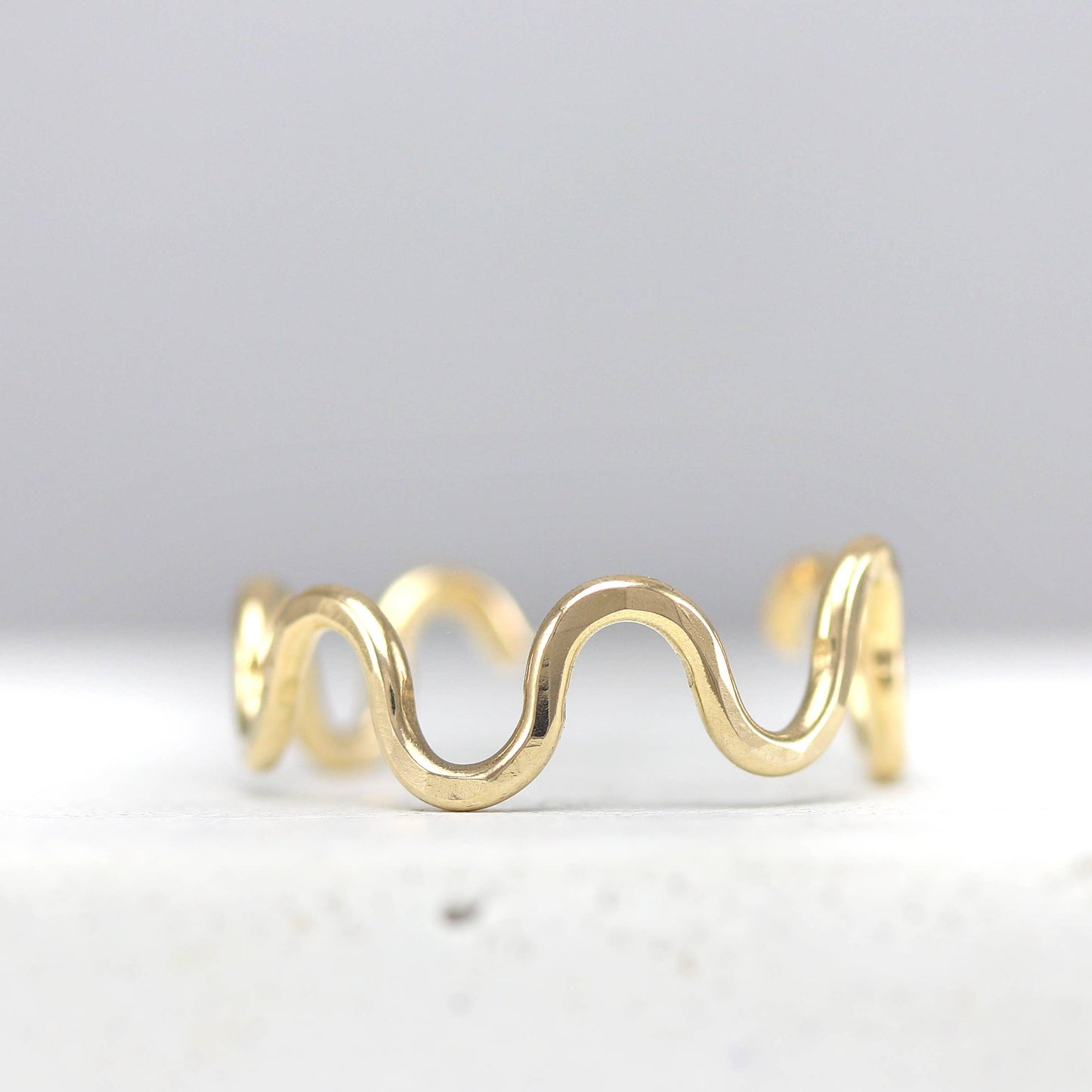 A thin gold ring with a continuous wavy, undulating design, photographed on a light gray background.