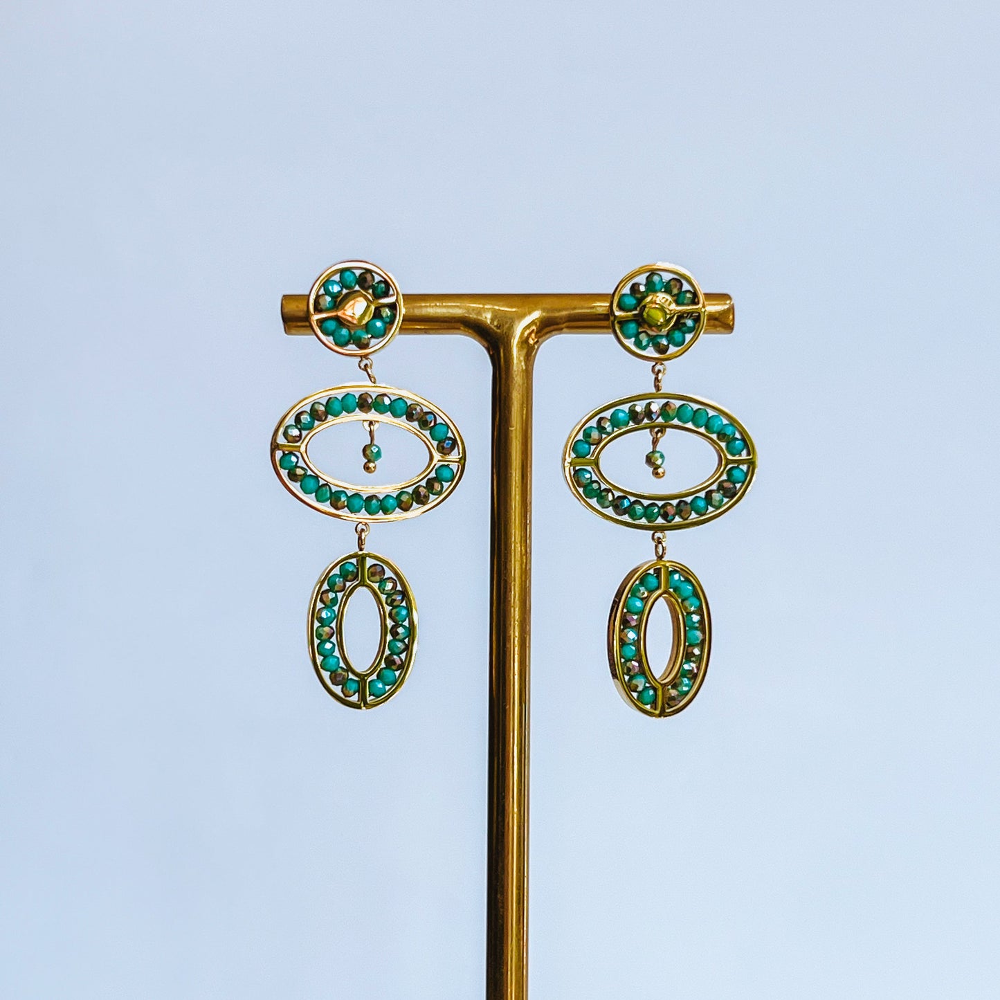 Abaco Earrings - Aquamarine Pair of gold earrings with green gemstones on a stand against a light background