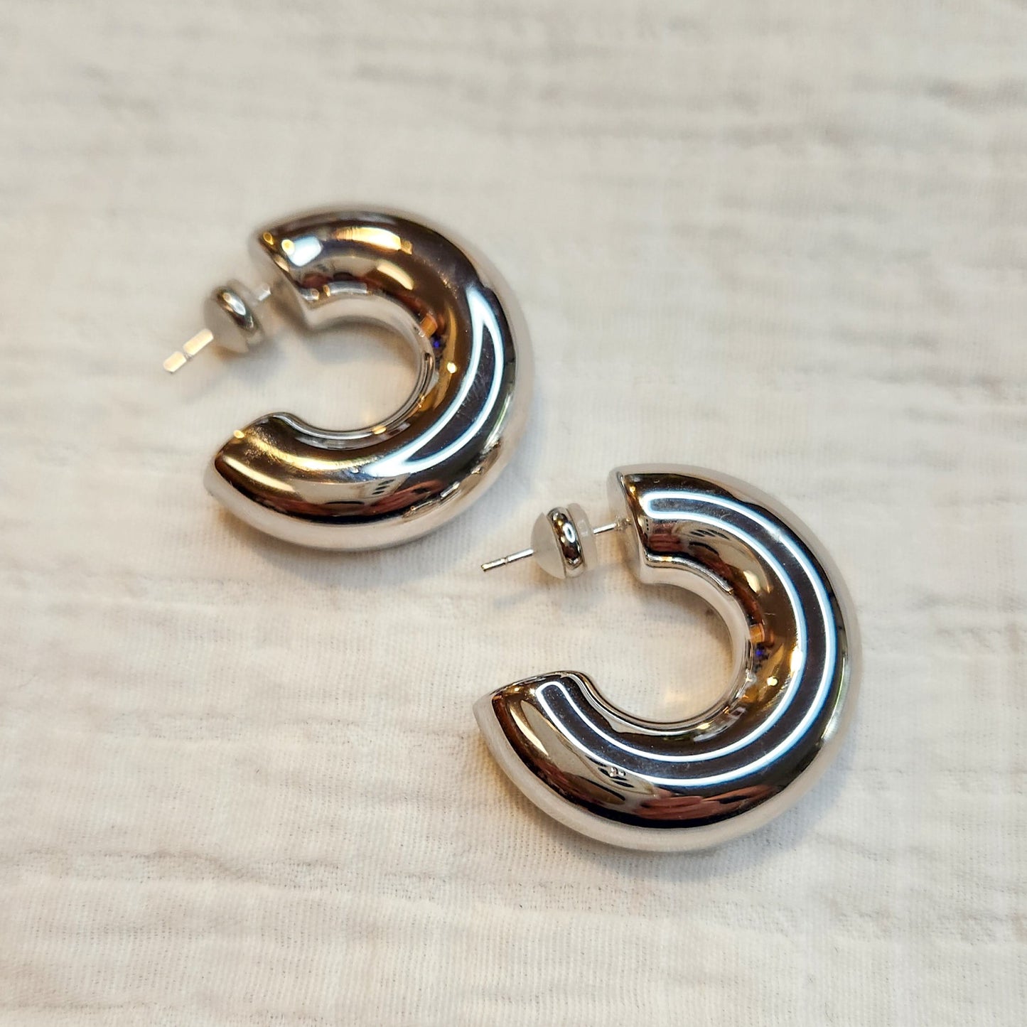 A pair of silver chunky large hoop earrings laid out on a textured fabric surface.