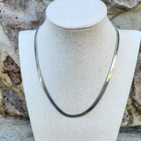 A silver-colored snake necklace displayed on a mannequin bust, set against a stone wall and floor background.