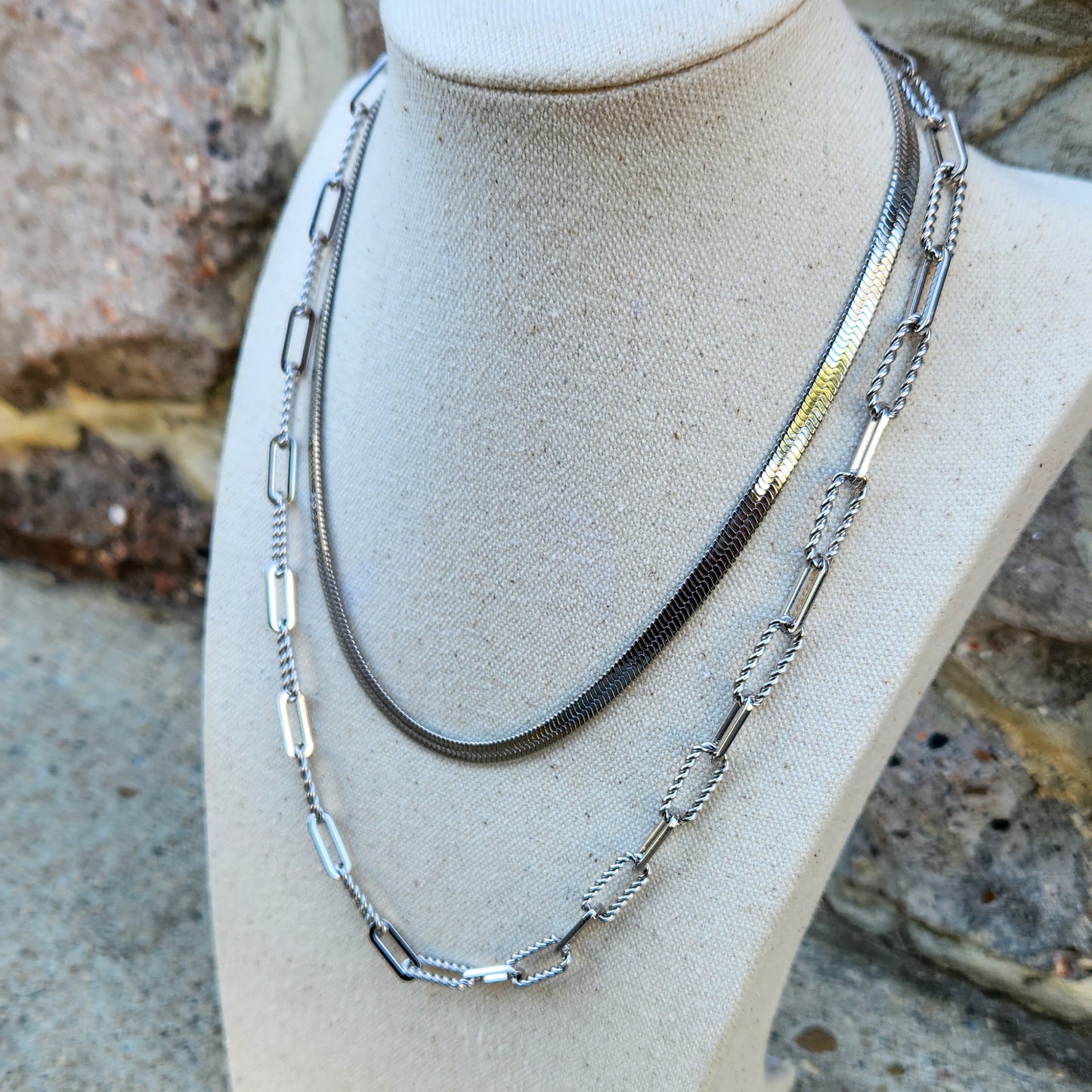 Two silver necklaces on a mannequin bust against a stone wall background