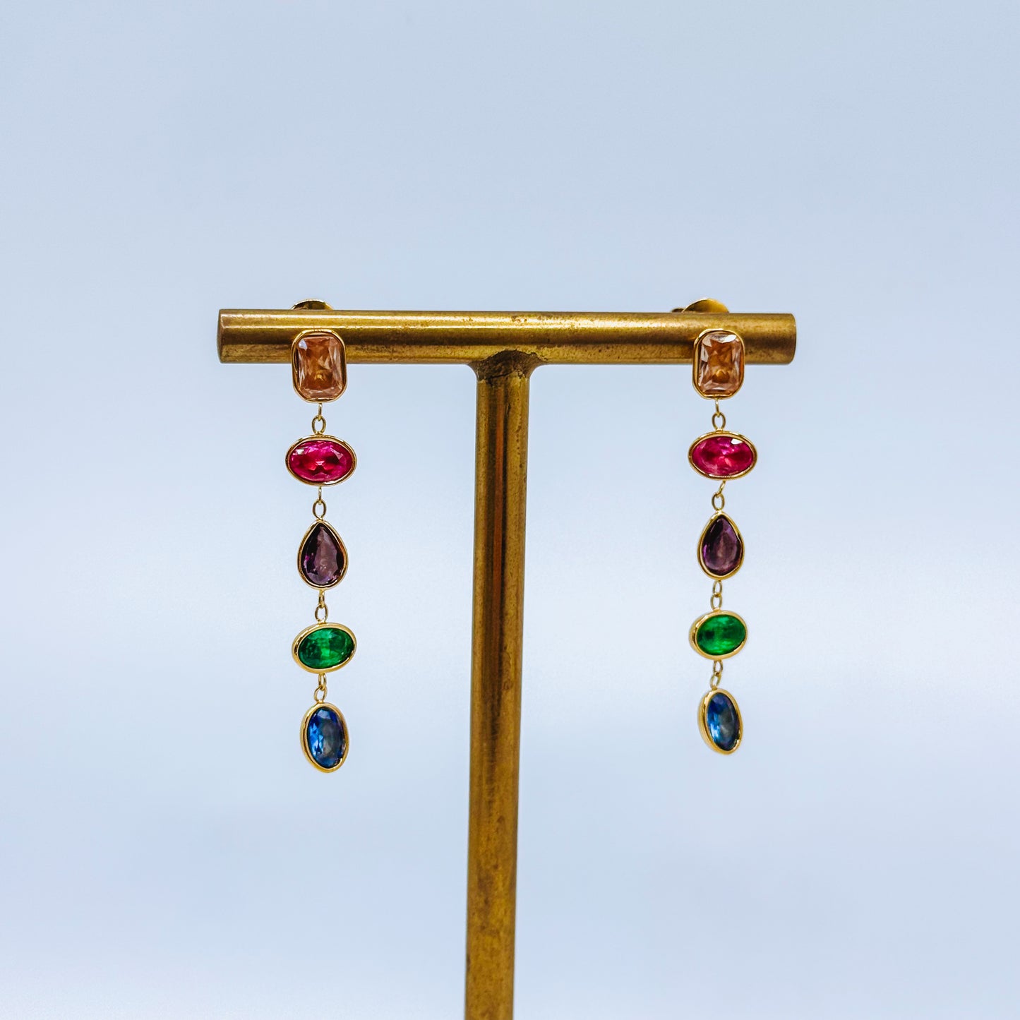 Pair of gold earrings with colorful gemstones on a gold stand against a light blue background