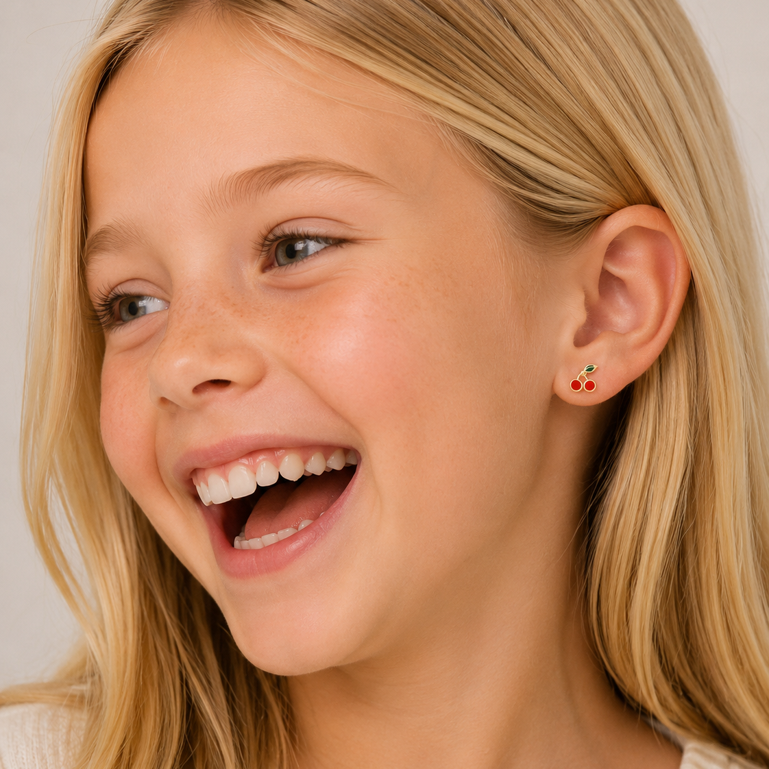 A close-up of a smiling young girl with blonde hair wearing a small red and green cherry flat back stud earring