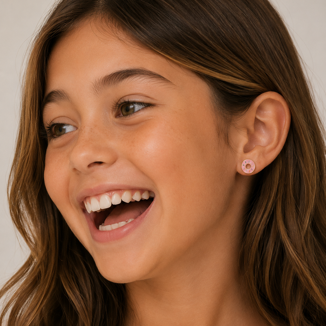 A close-up of a smiling young girl with brown hair wearing a small pink donut stud gold flat back earring 