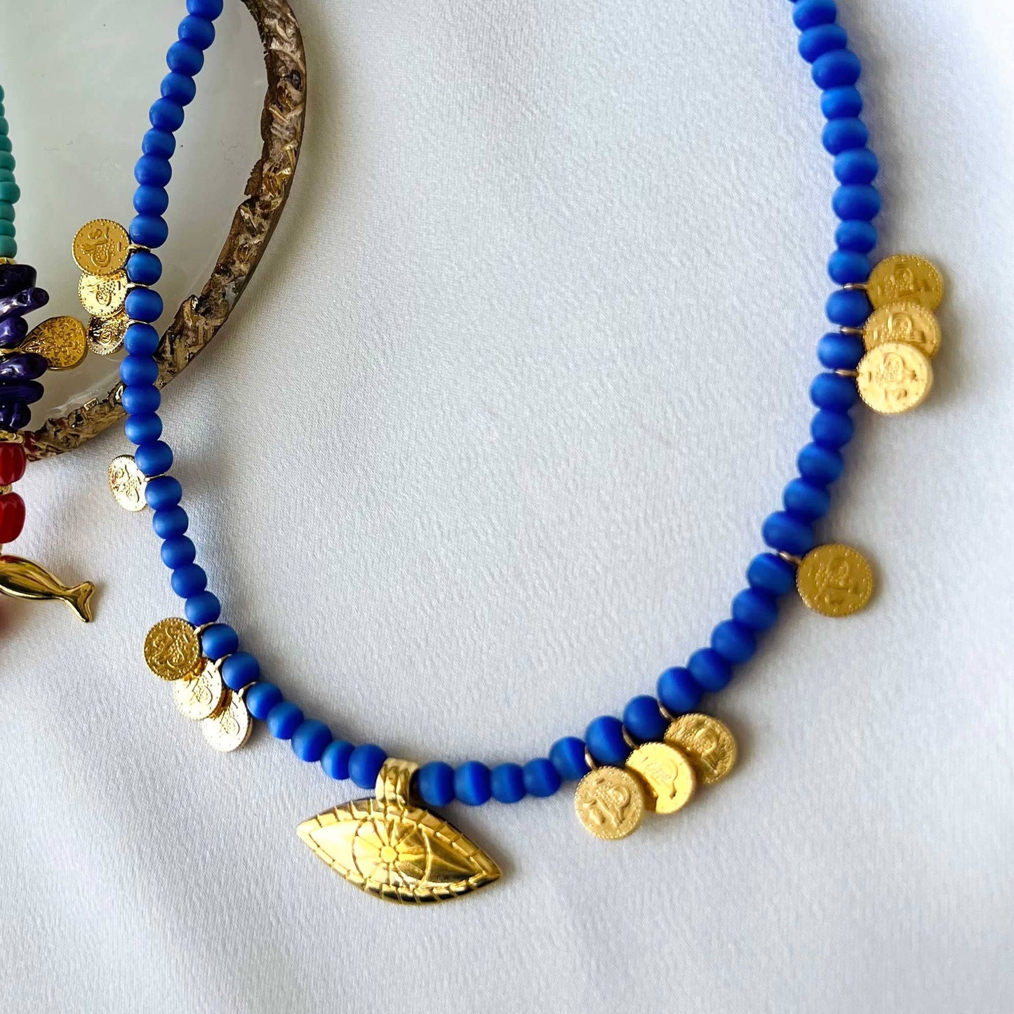 A blue beaded necklace with goldplated copper parts, including a goldplated eye-shaped charm, displayed on a white surface.
