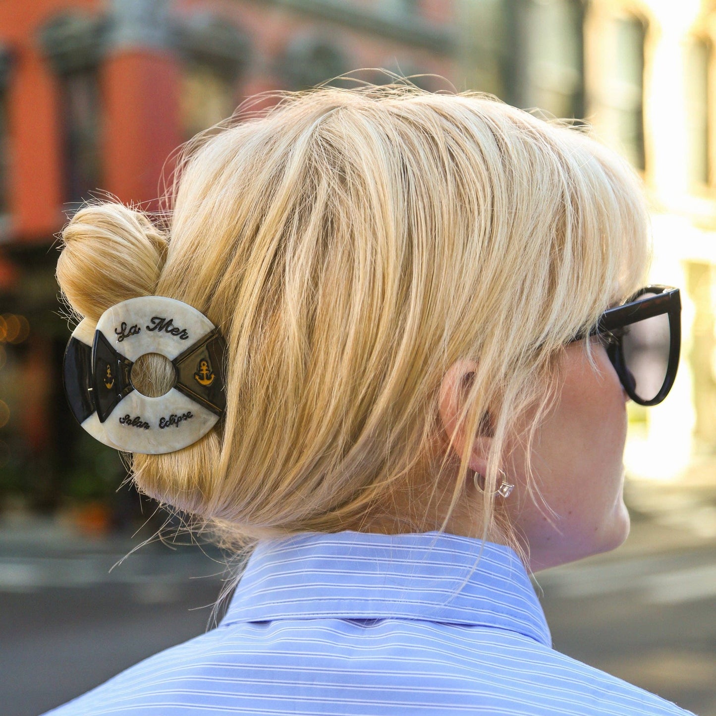 Person with blonde hair styled in a bun with a Hand- painted black and white life buoy claw hair clip with 'La Mar' branding also wearing sunglasses and a blue shirt.