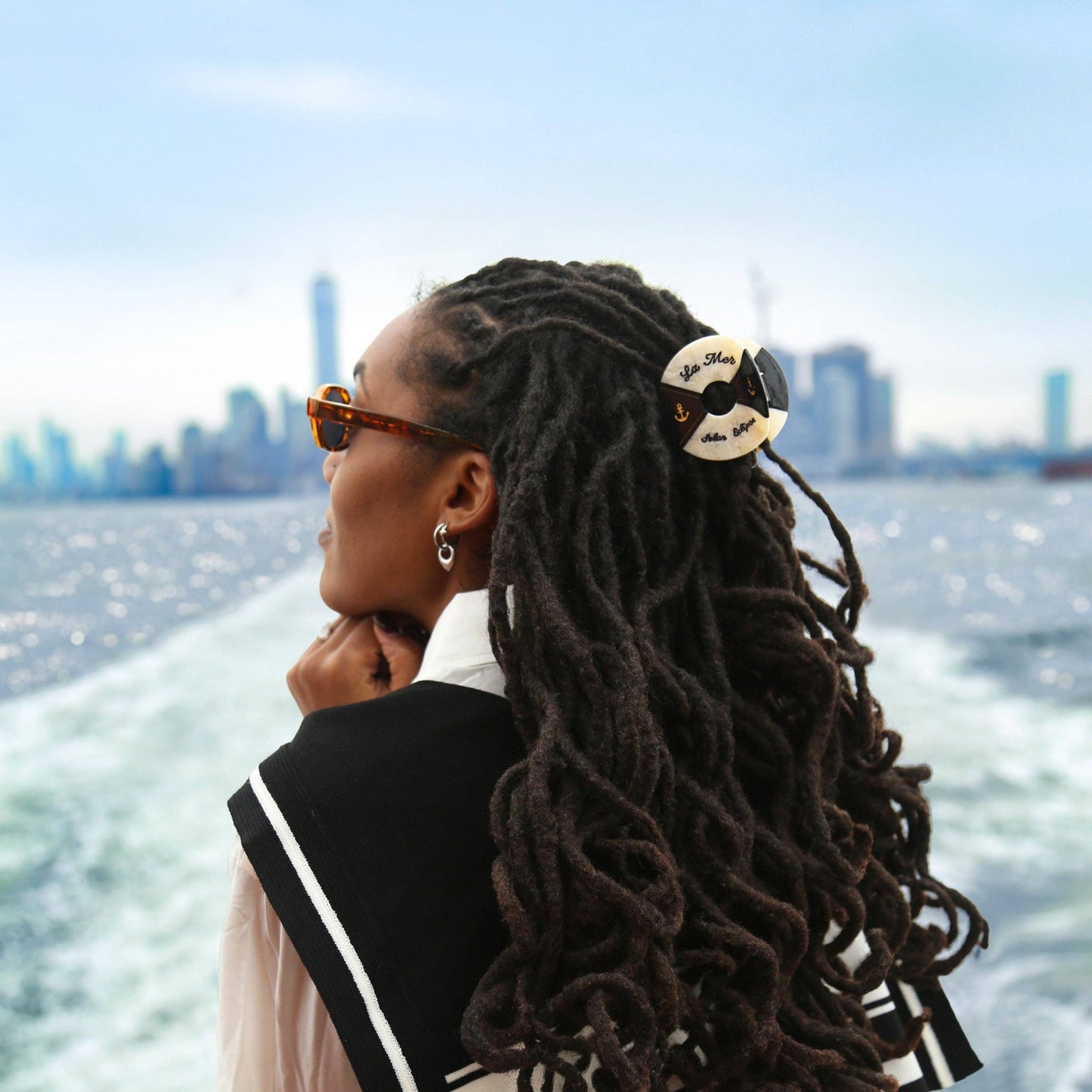 Woman with styled hair with a Hand- painted black and white life buoy claw hair clip with 'La Mar' branding sitting on a boat with a city skyline in the background