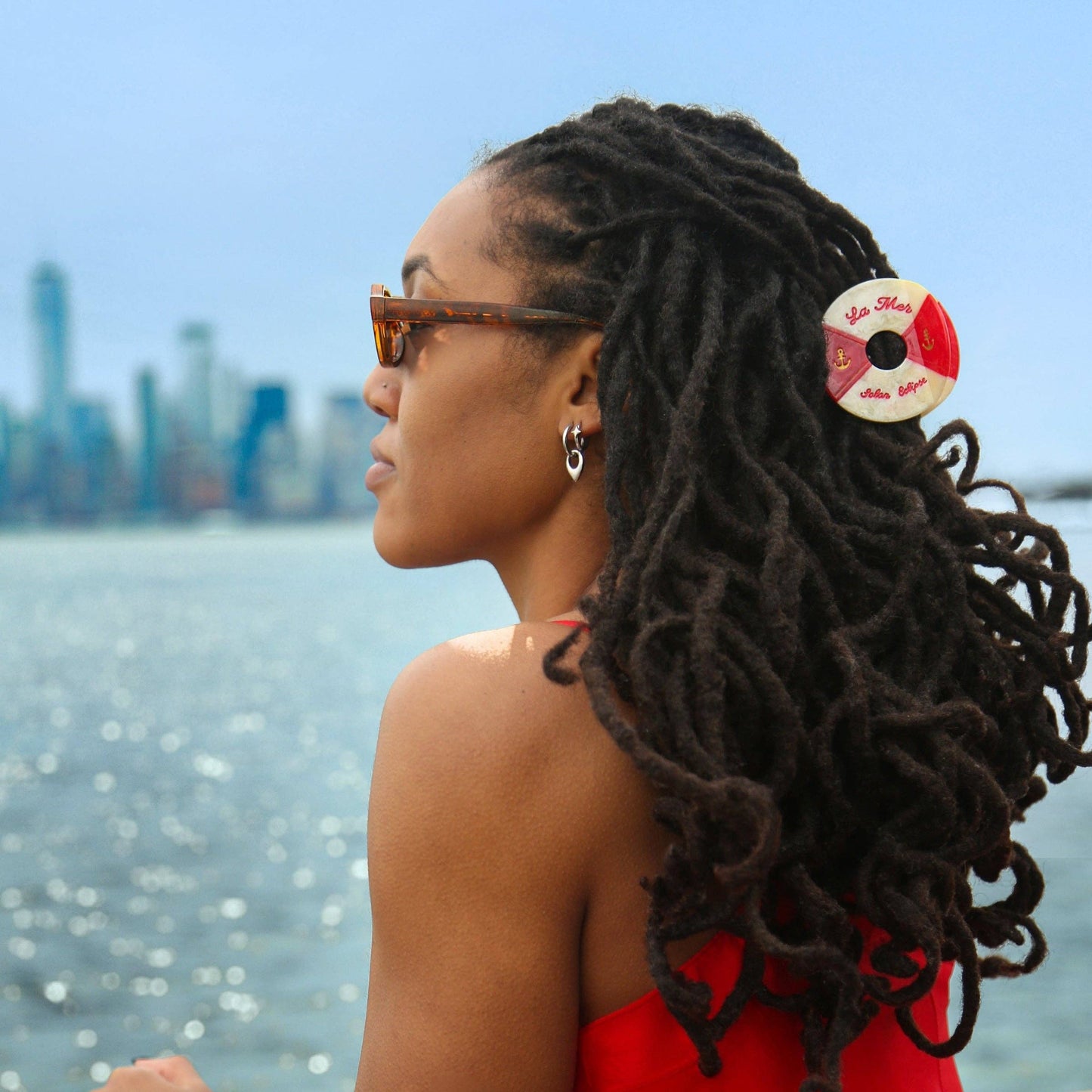 Woman with styled hair with a Hand- painted red and white life buoy claw hair clip with 'La Mar' branding sitting by a waterfront with a city skyline in the background