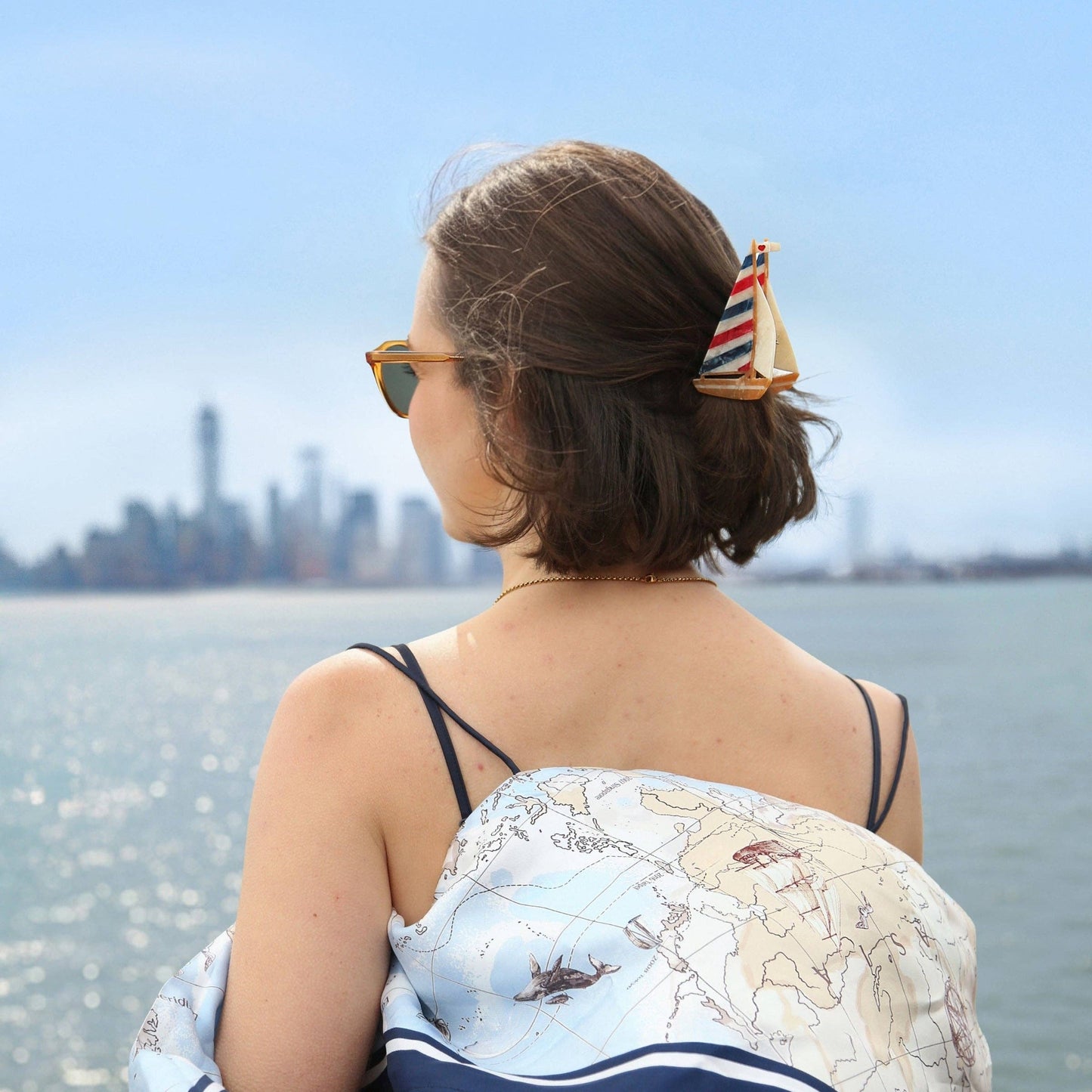 Woman with a map-patterned scarf over her shoulder and hair styled with a Hand-painted Sailboat Claw Hair Clip, looking at a city skyline from a waterfront.