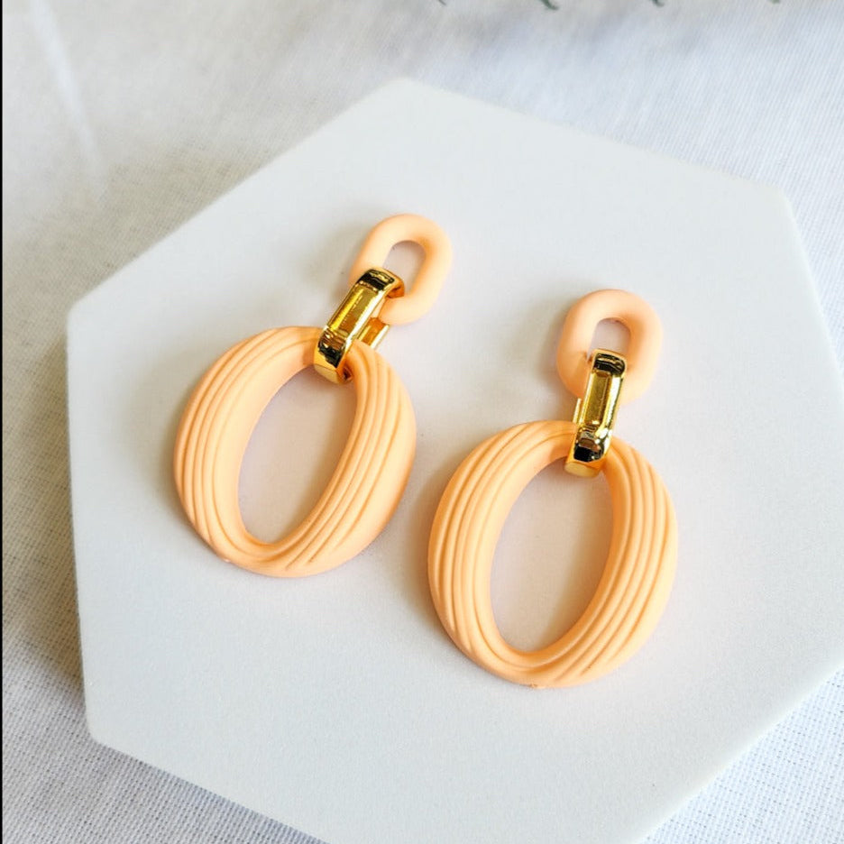 A pair of peach-colored, loop-shaped Mavra earrings with gold accents on the hooks, displayed on a white surface.