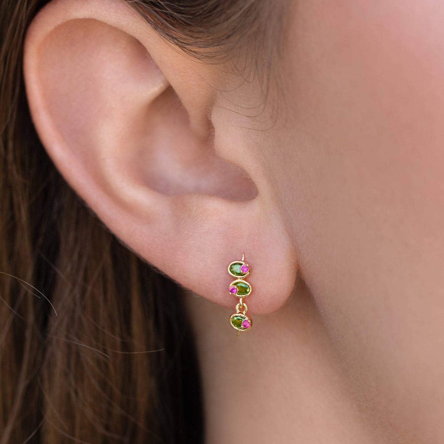 Mini Olive Studs: Gold Close-up of an ear wearing a gold earring with green and pink olive shaped gemstones.