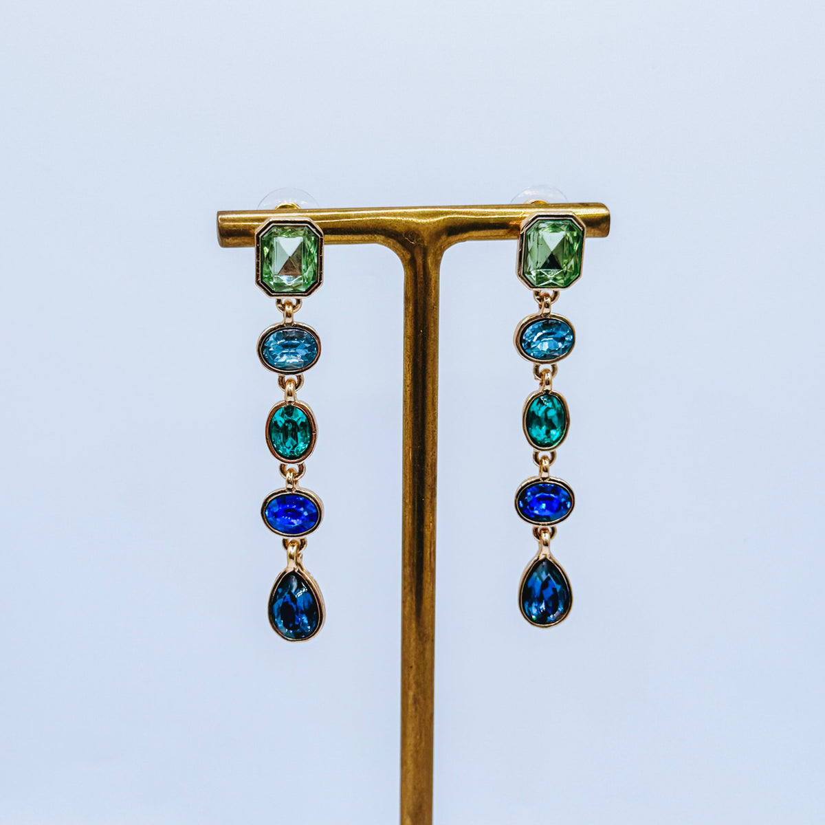 A pair of gold-tone drop earrings with green rectangular top stones and cascading blue and emerald-cut stones, displayed on a gold T-stand against a light blue background.