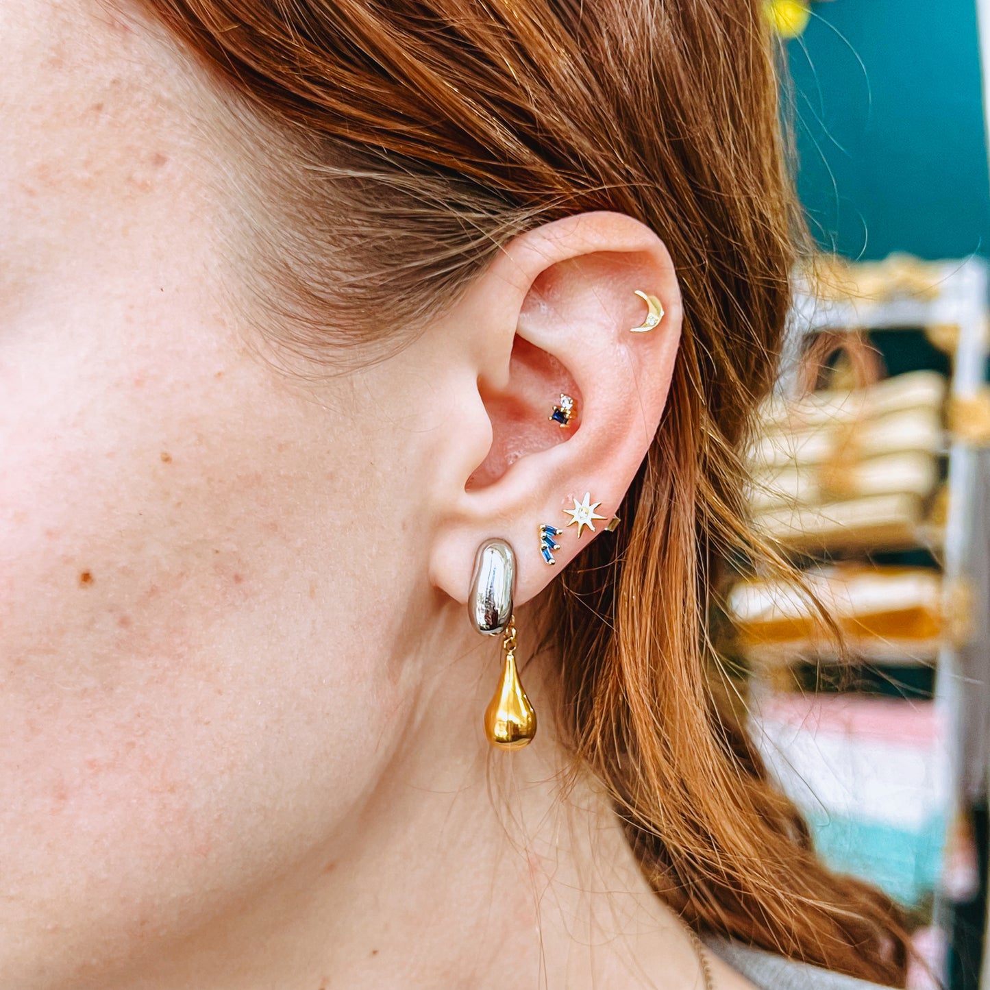 Close-up of an ear with multiple earrings including the gold and silver Tear Drop Mixed Metals Earrings against a blurred background
