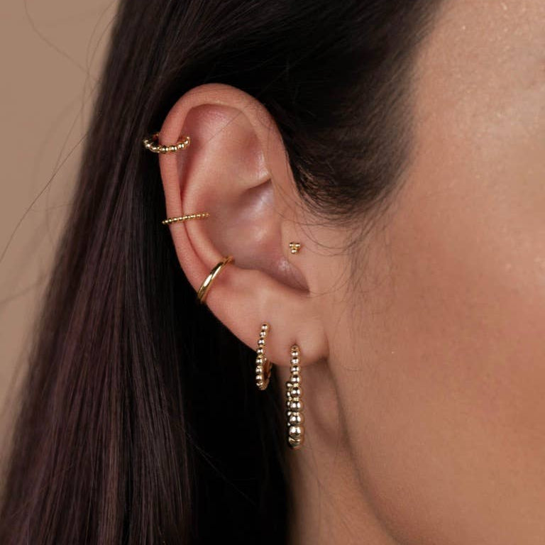 Tiny Gold Earrings - Ball Hoop Close-up of an ear with gold earrings and a ring against a neutral background