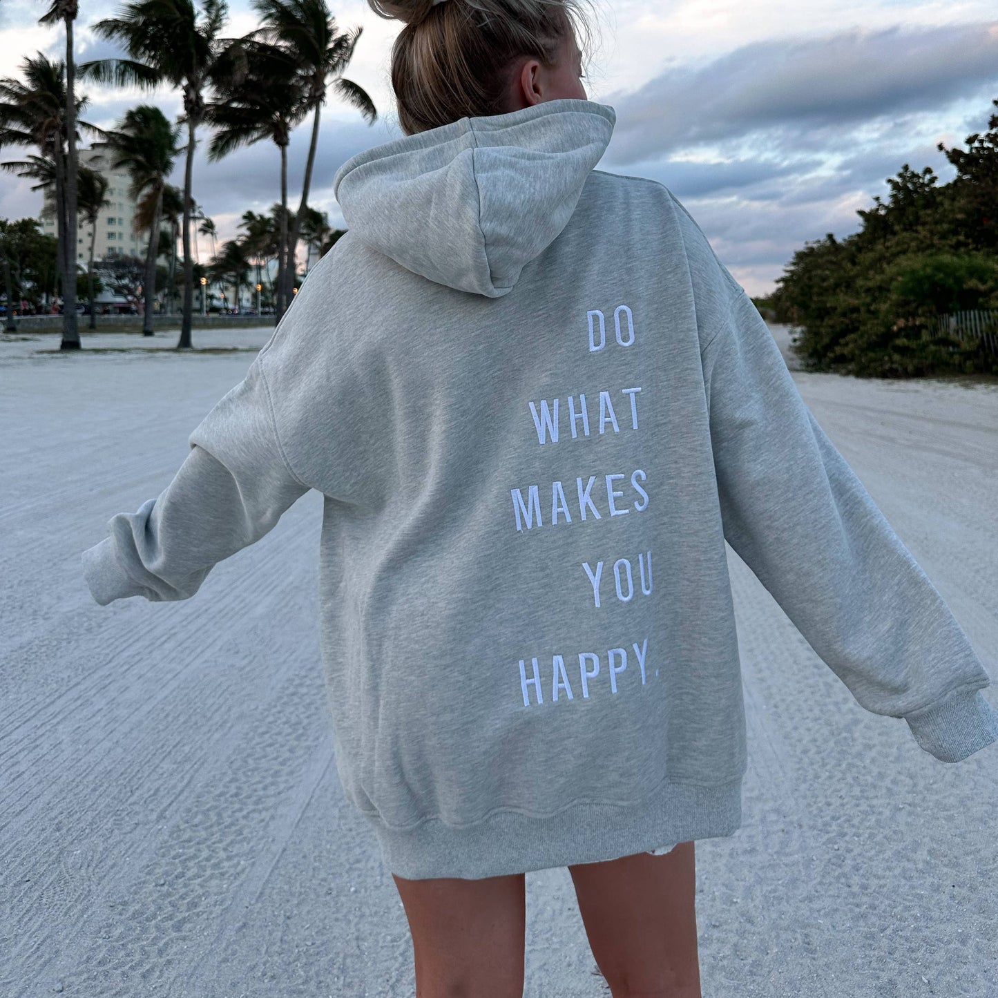 Do What Makes You Happy Embroidered Hoodie - Gray Person wearing a gray hoodie with 'Do What Makes You Happy' text on a beach.
