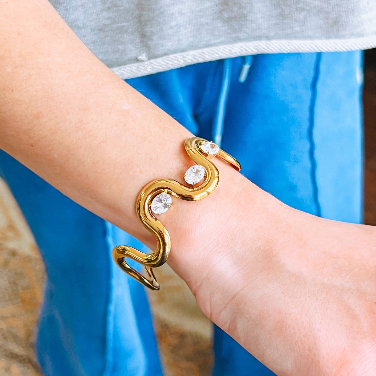 Gold wave-shaped bracelet with three clear stones on a wrist against a blue fabric background